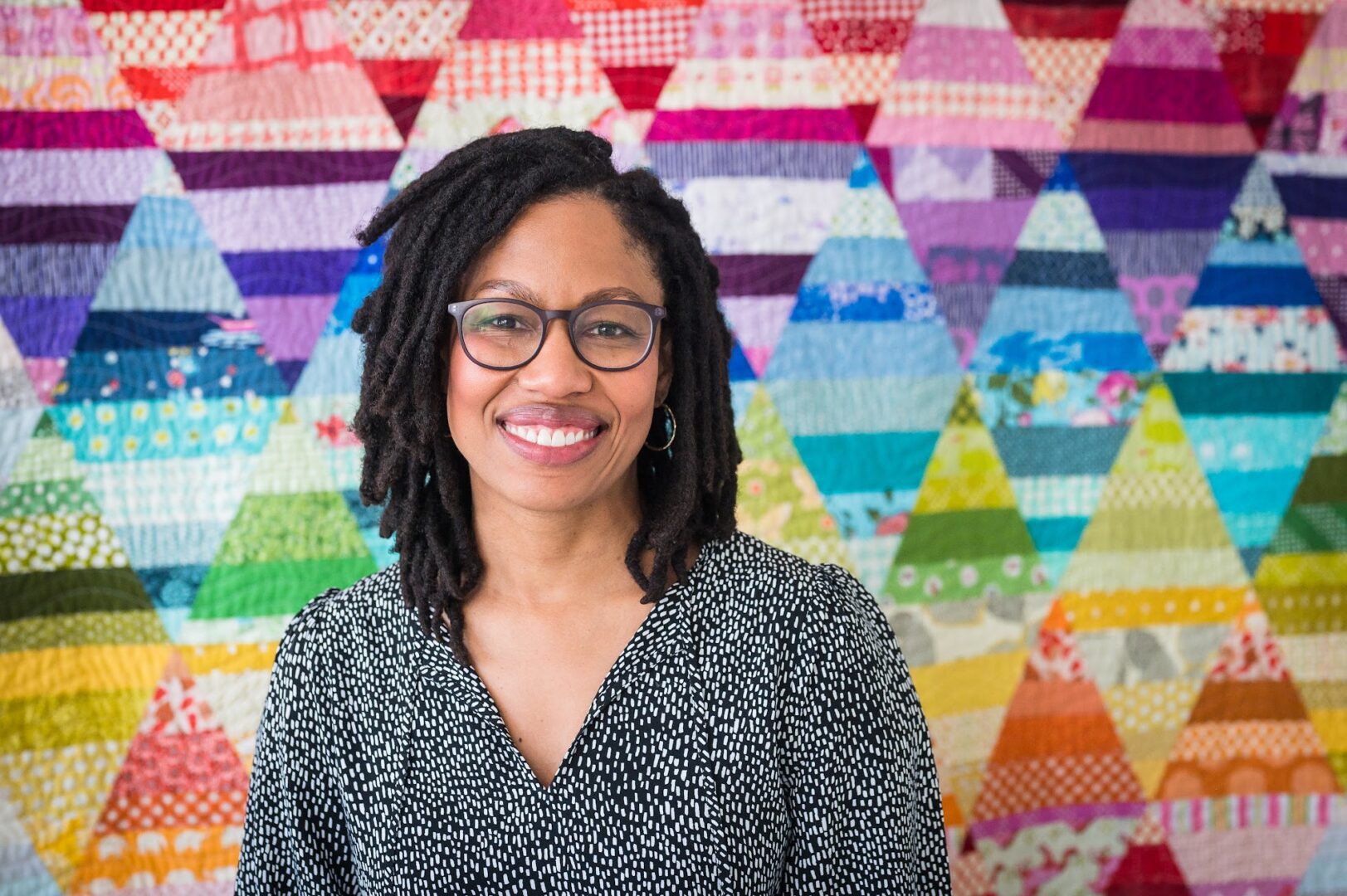A woman with glasses standing in front of a colorful quilt.