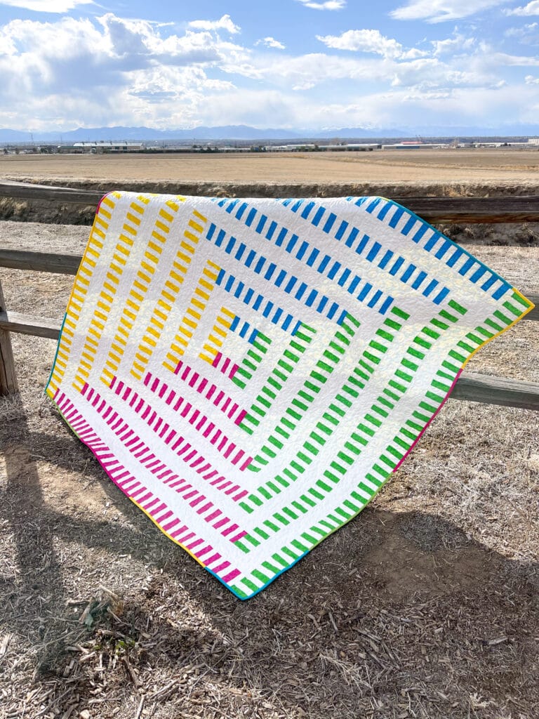 A colorful quilt hanging on a fence in a field.