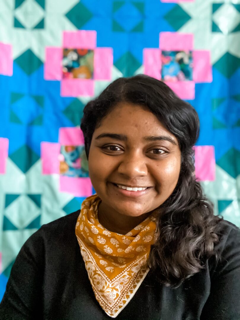 A young woman smiling in front of a colorful quilt.
