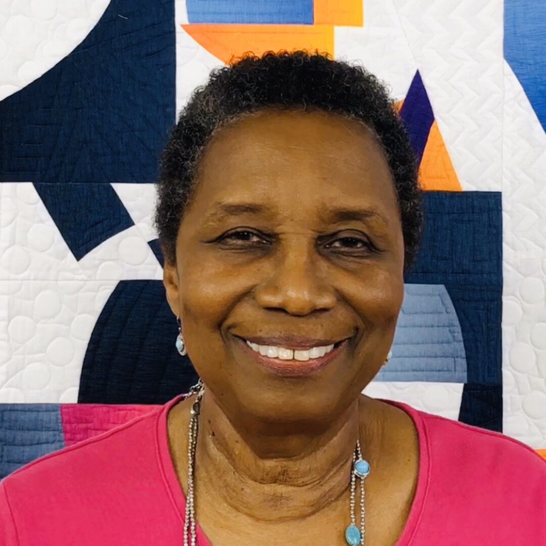 An older woman smiling in front of a colorful quilt.