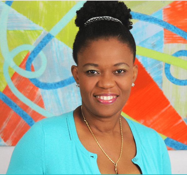 A woman in a blue shirt smiling in front of a colorful painting.