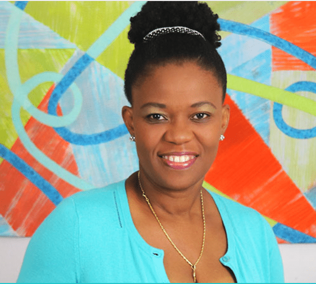 A woman in a blue shirt smiling in front of a colorful painting.