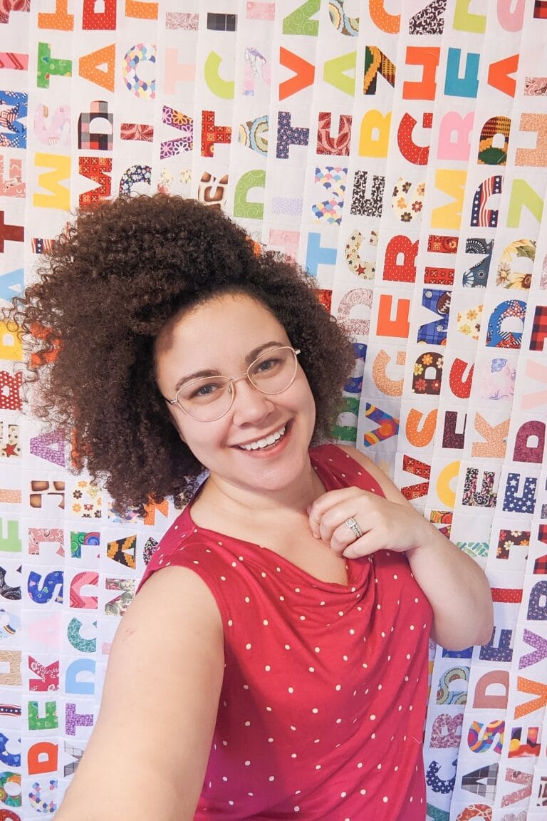 A woman in glasses is taking a selfie in front of a colorful wall of letters.
