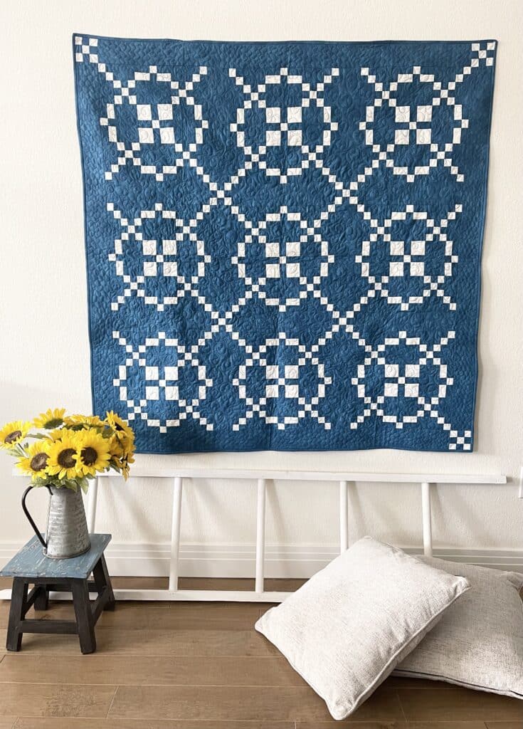 A blue quilt hanging on a wall next to a vase of sunflowers.