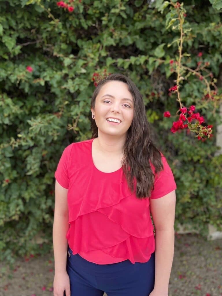 A young woman in a red top standing in front of a bush.