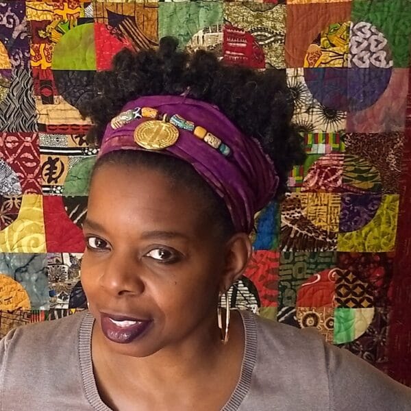 A woman wearing a purple headband in front of a colorful quilt.