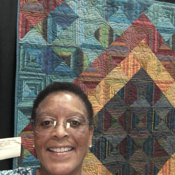 A woman with glasses smiling in front of a quilt.