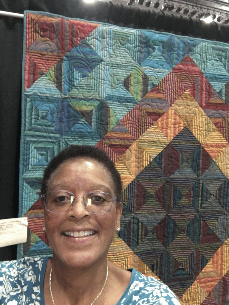 A woman with glasses smiling in front of a quilt.