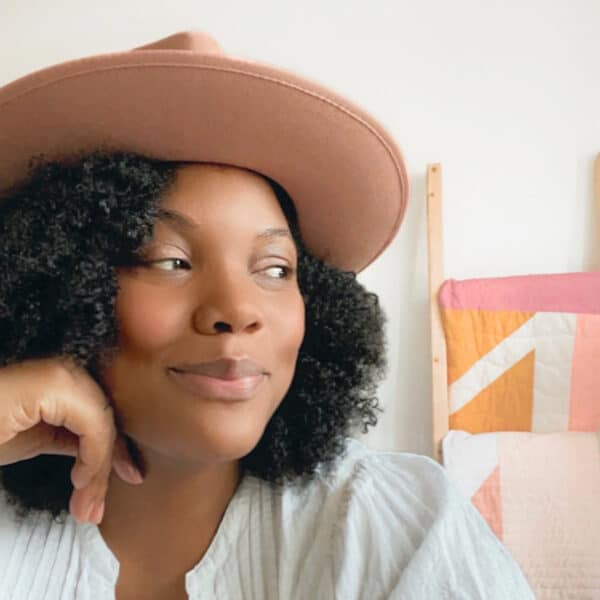 A woman wearing a hat in front of a quilt.