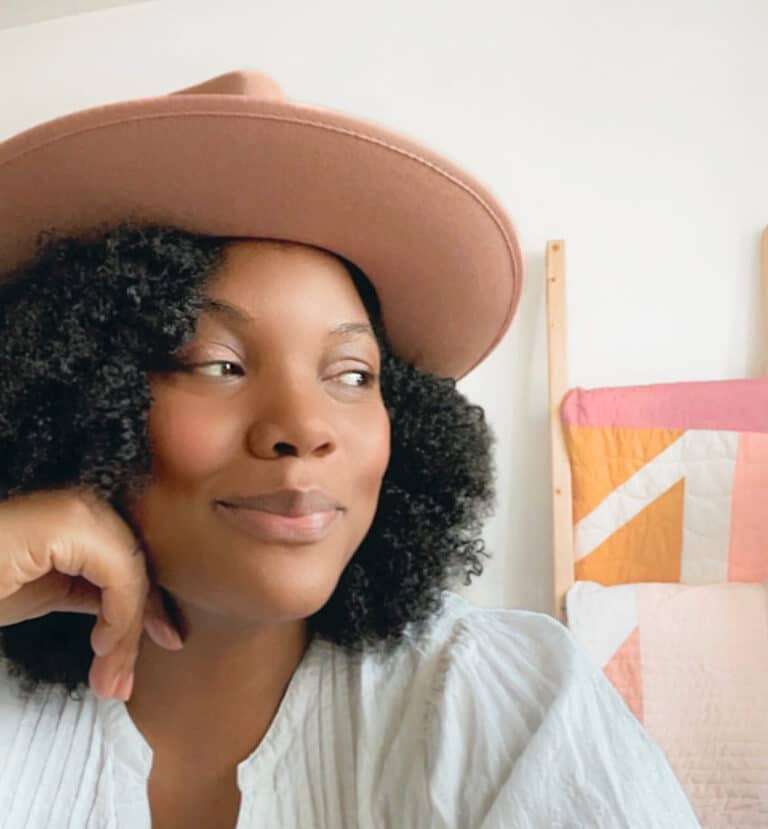 A woman wearing a hat in front of a quilt.