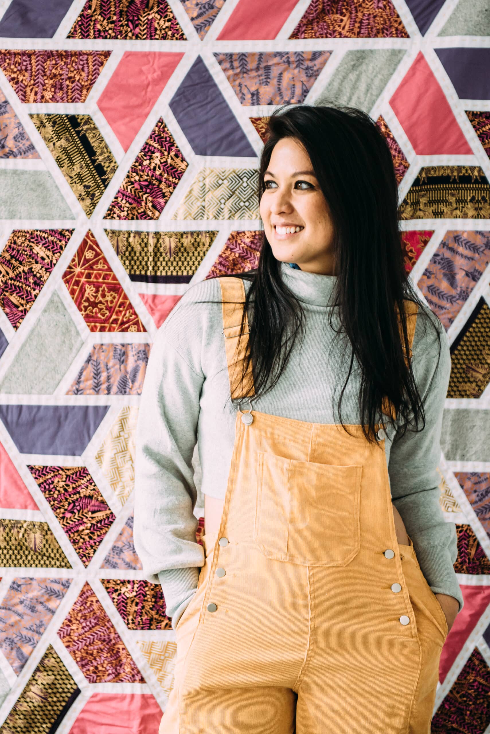 A woman in overalls standing in front of a quilted wall.